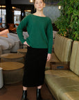 A woman standing in a lobby setting, wearing a green knit sweater and a black skirt, with various seating options and plants in the background.
