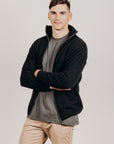 Man wearing a black  zip jacket over a gray shirt with beige pants on a plain background