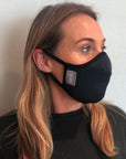 Woman wearing a black travel mask with adjustable elastic straps, made from merino soft & comfort next to skin, with a label showing 'Made in New Zealand', and highlighting features such as 100% breathable, adjustable elastic straps, and a machine washable filter.