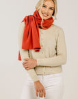 Woman wearing a beige sweater and white pants with a red scarf on a plain background