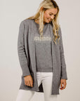 Woman wearing a gray cardigan over a white top with a subtle pattern, standing against a plain background.