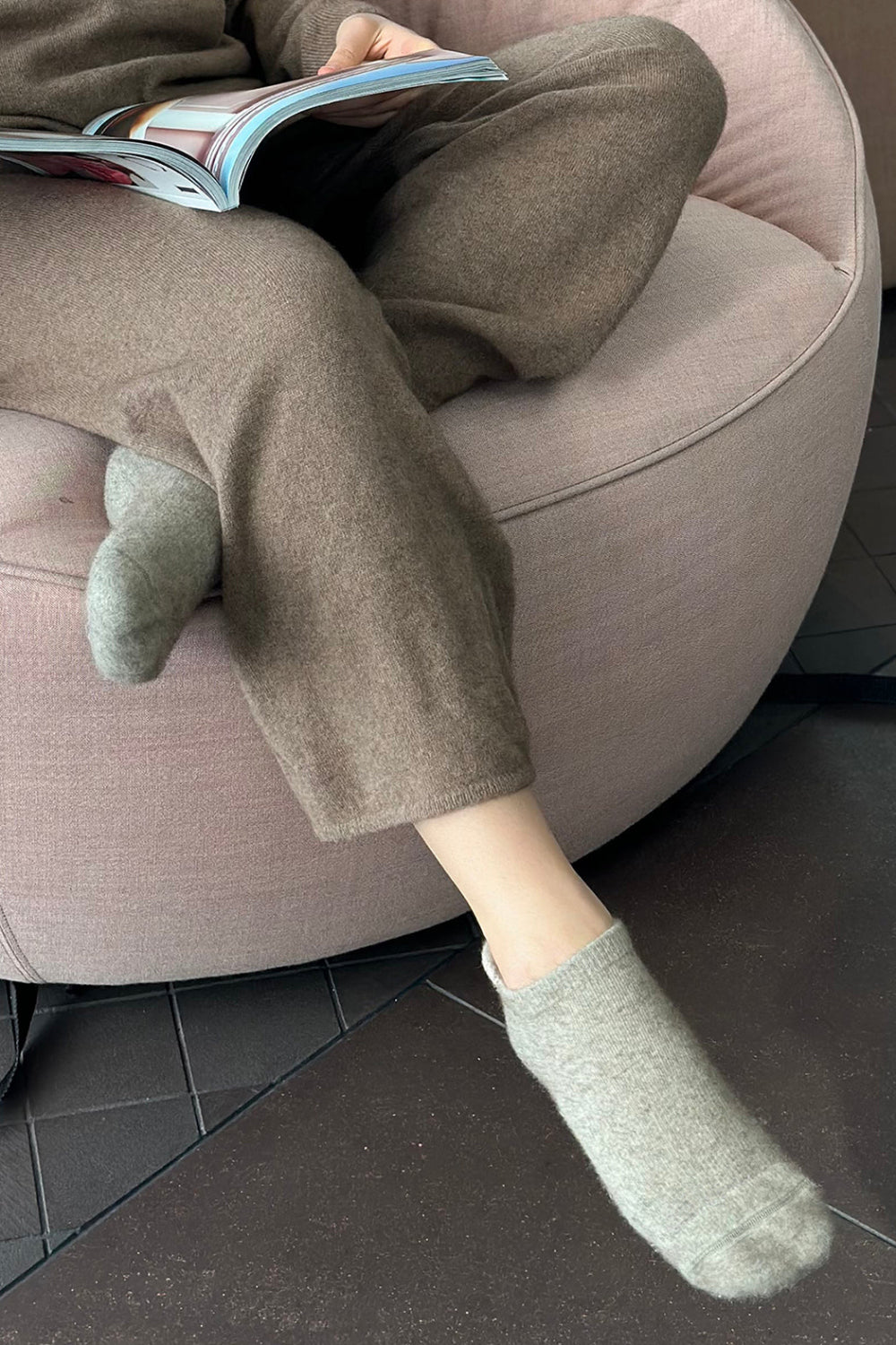 Person wearing brown slipper socks with the ankle area exposed, seated in a pink round chair reading a book.