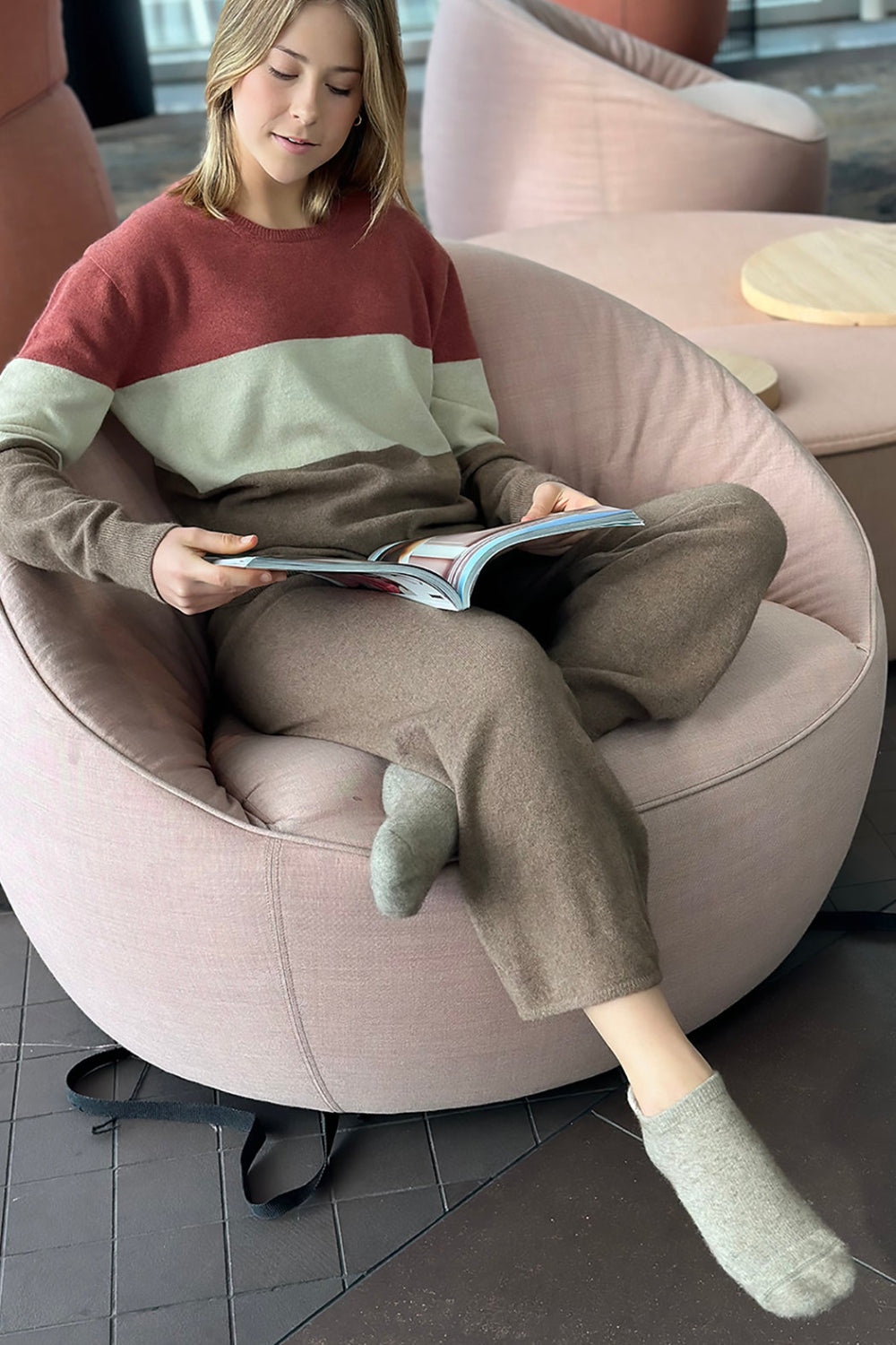 Person wearing brown slipper socks with the ankle area exposed, seated in a pink round chair reading a book.