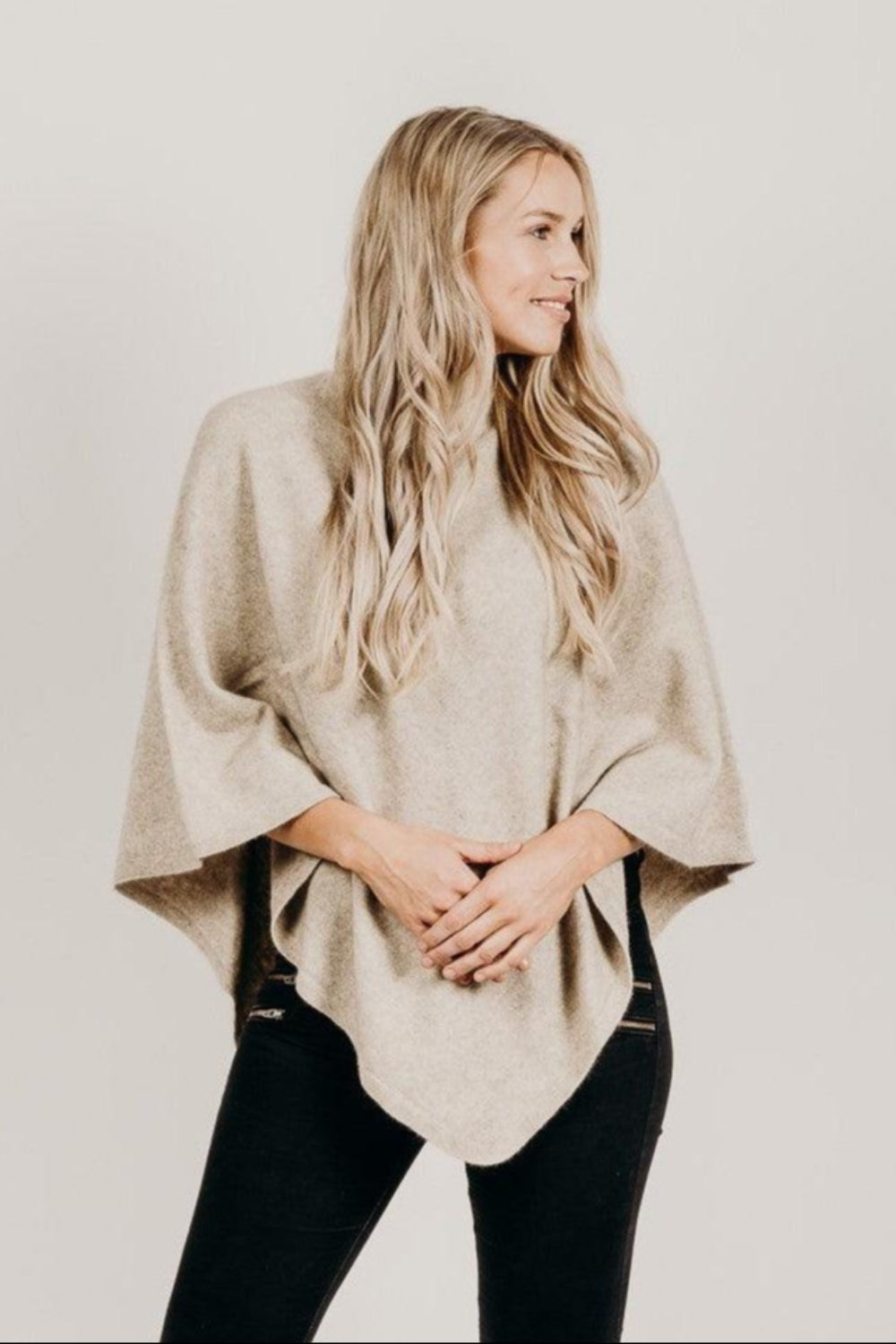 A woman wearing a beige Merinosilk poncho over a black top, paired with black pants. The poncho has a flowing design and she has long blonde hair.