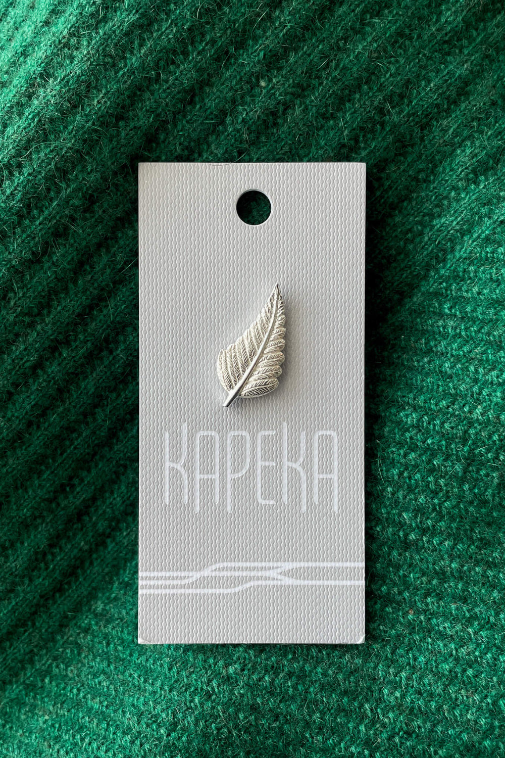 Silver leaf-shaped brooch on a Kapeka card against a green textured background