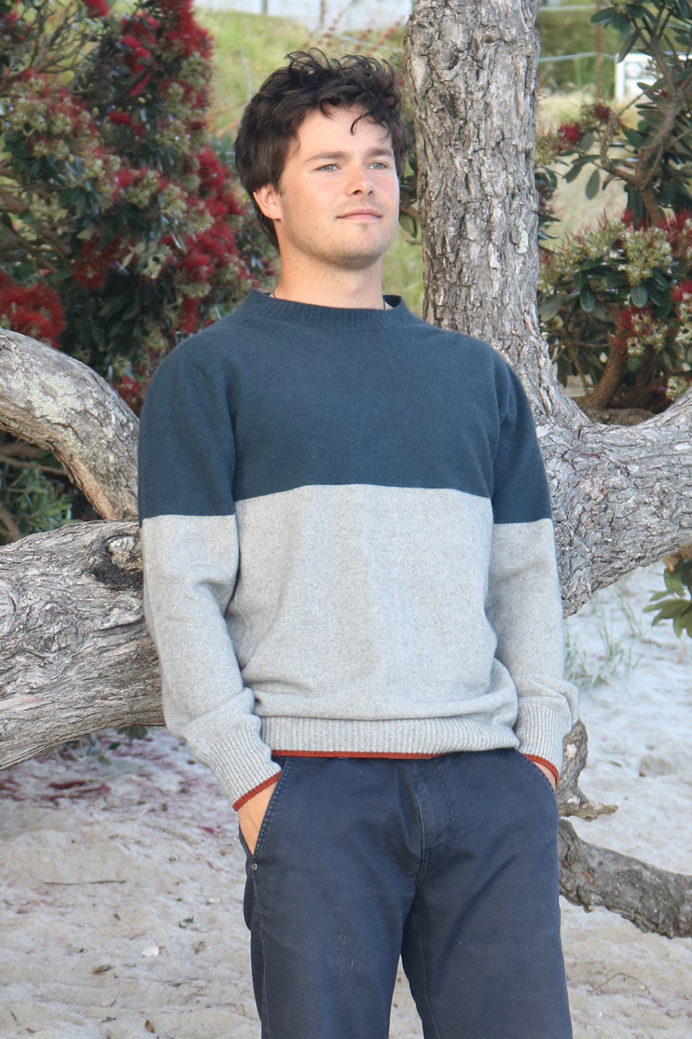 A person standing by a tree, wearing a striped Merinosilk Traverse Sweater in navy and gray, with a linked crew neckline and ribbed details on the shoulders.