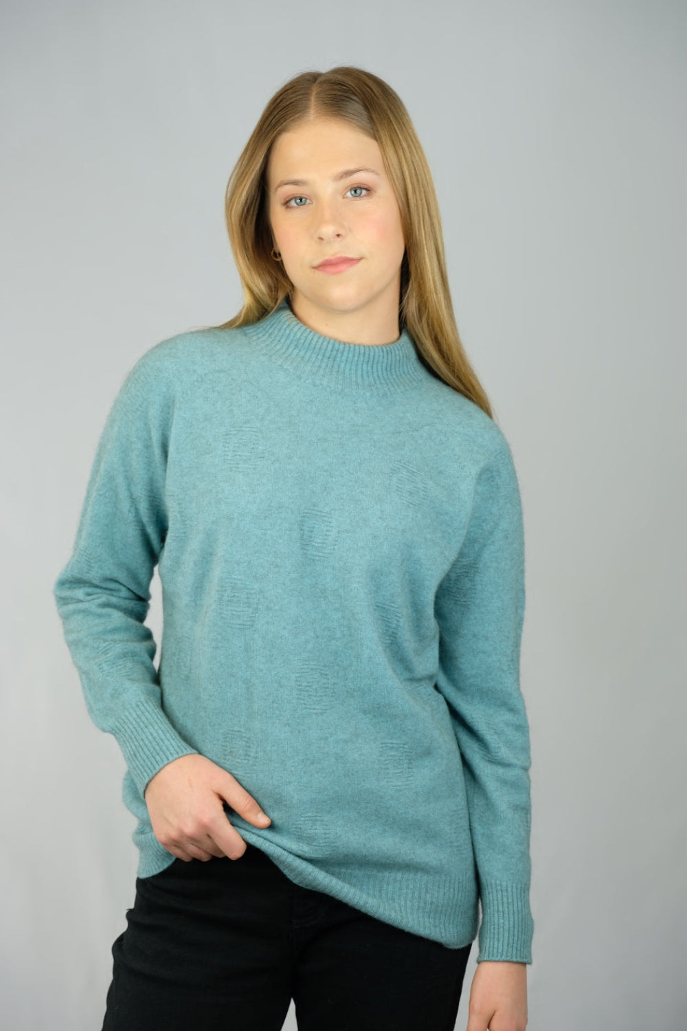 A woman wearing a light green turtle neck sweater with ribbed cuffs and a crew neckline, paired with black pants.