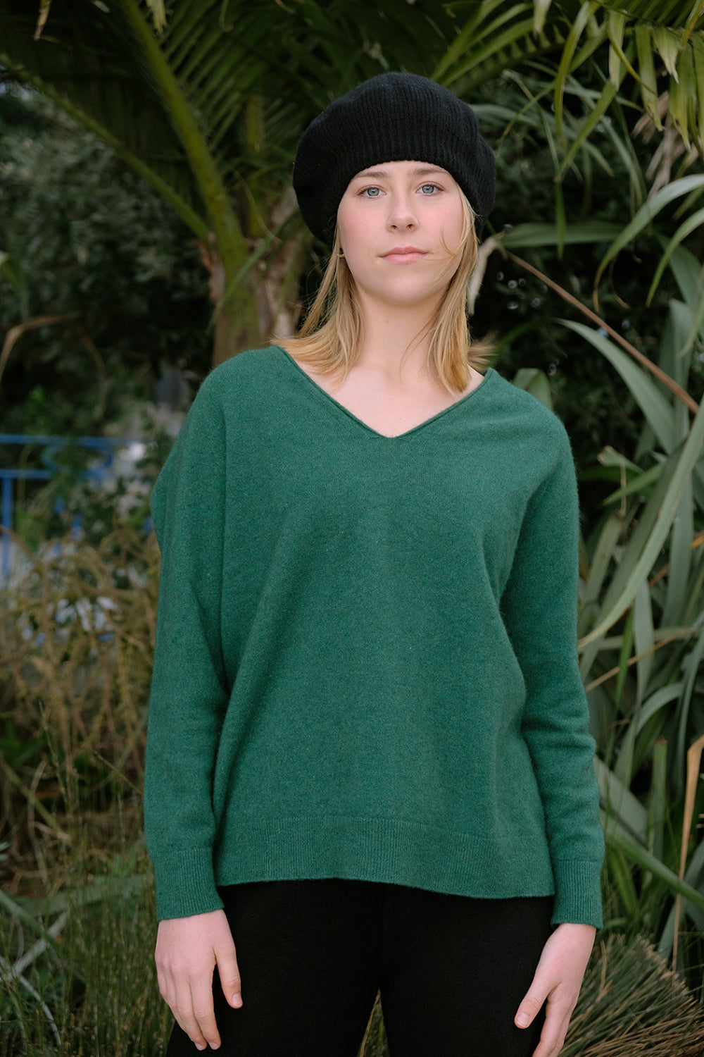 Person wearing a green sweater and black beanie standing outdoors with greenery in the background