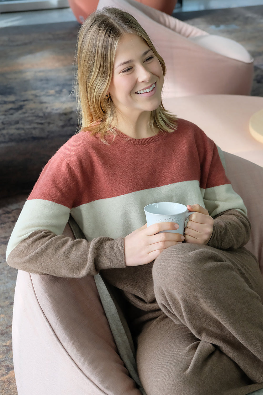A woman sitting in a cozy position wearing a brown Merinosilk Queenstown Sweater with red and white stripes, holding a coffee cup, and smiling.