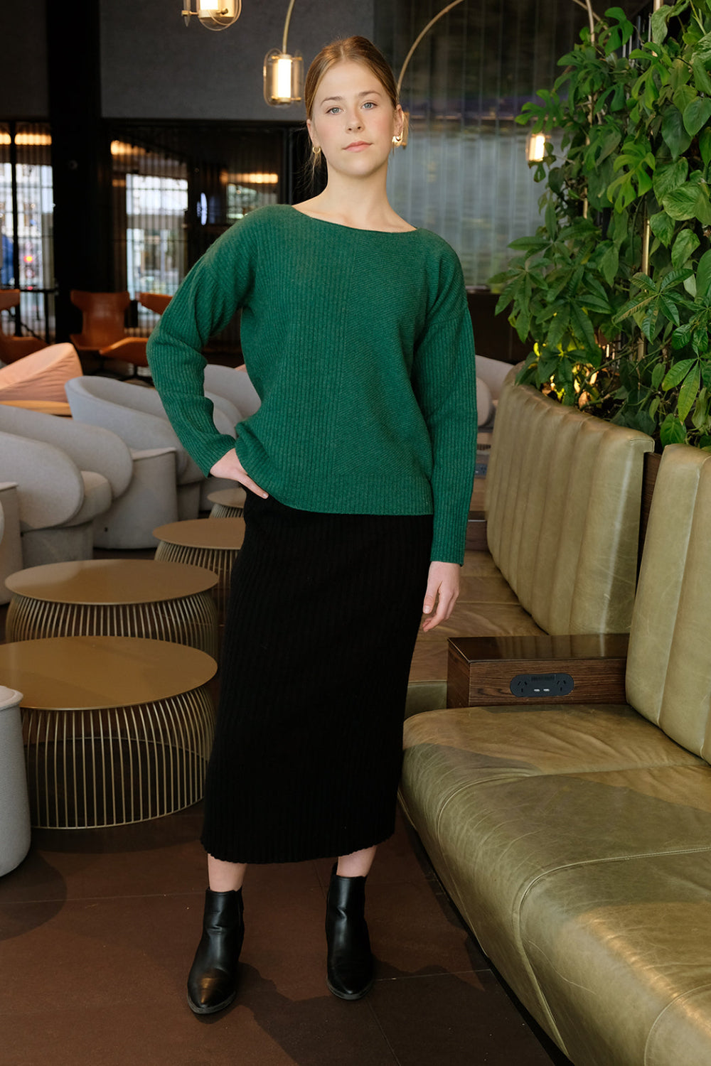 A woman standing in a lobby setting, wearing a green knit sweater and a black skirt, with various seating options and plants in the background.