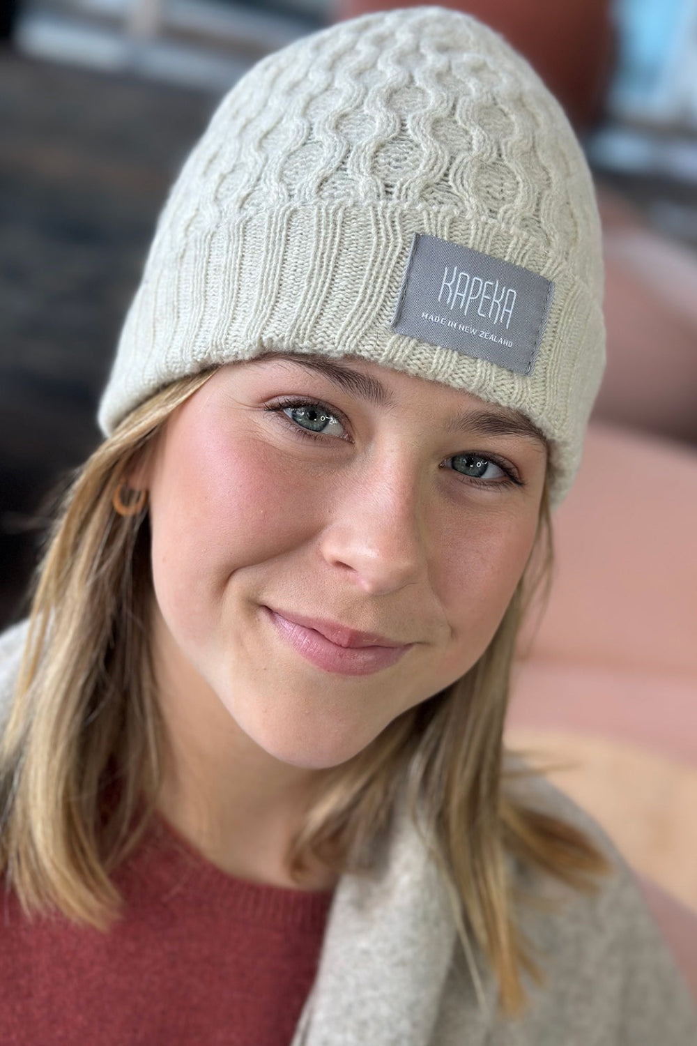 A person wearing a beige cable knit beanie with the 'Kapeka' brand label visible on the front.