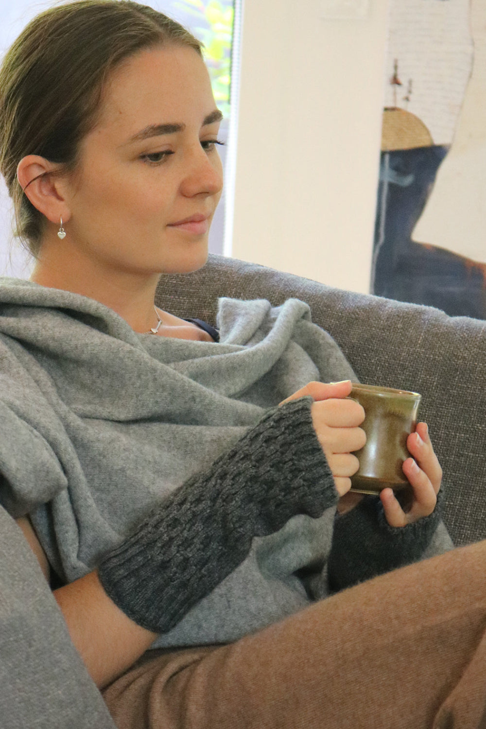 A person sitting on a sofa wearing a gray Merinosilk Tekapo wrist warmer while holding a cup.