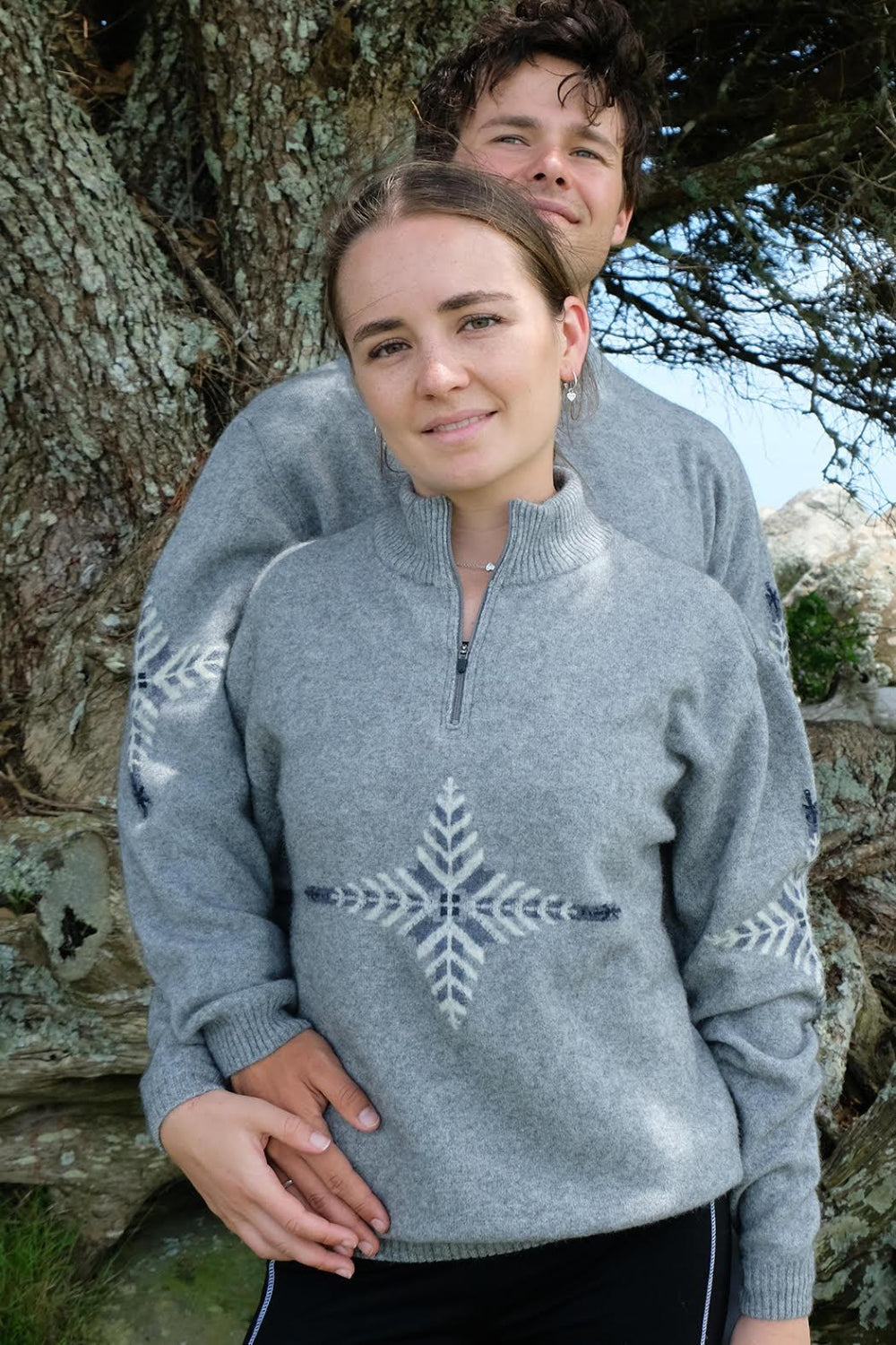 Two people wearing gray sweaters with a snowflake design, standing outdoors near a tree.