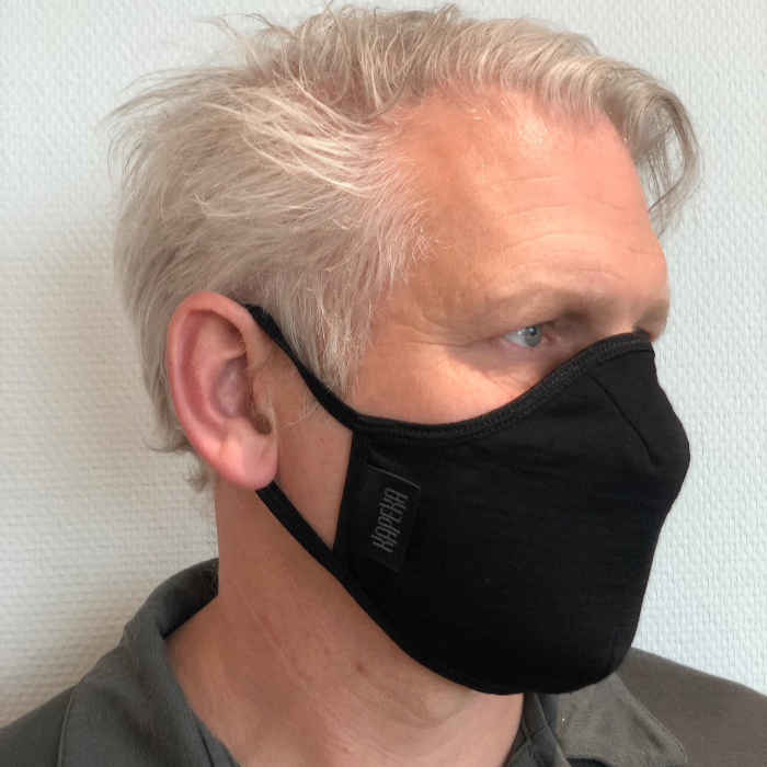 Man wearing a black travel mask with adjustable elastic straps, made from merino soft &amp; comfort next to skin, with a label showing &#39;Made in New Zealand&#39;, and highlighting features such as 100% breathable, adjustable elastic straps, and a machine washable filter.