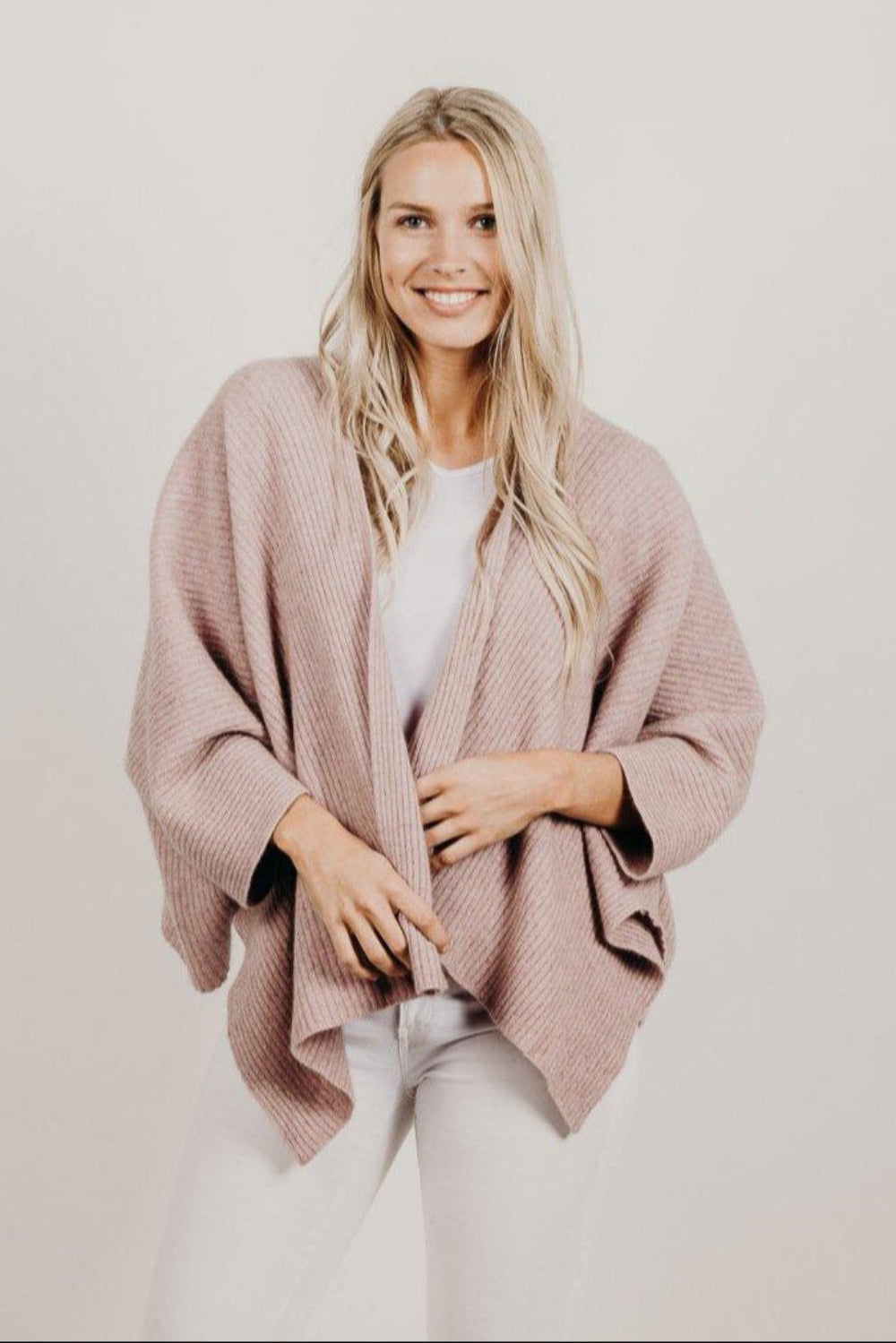 Woman wearing a pink poncho over a white top and pants on a plain background