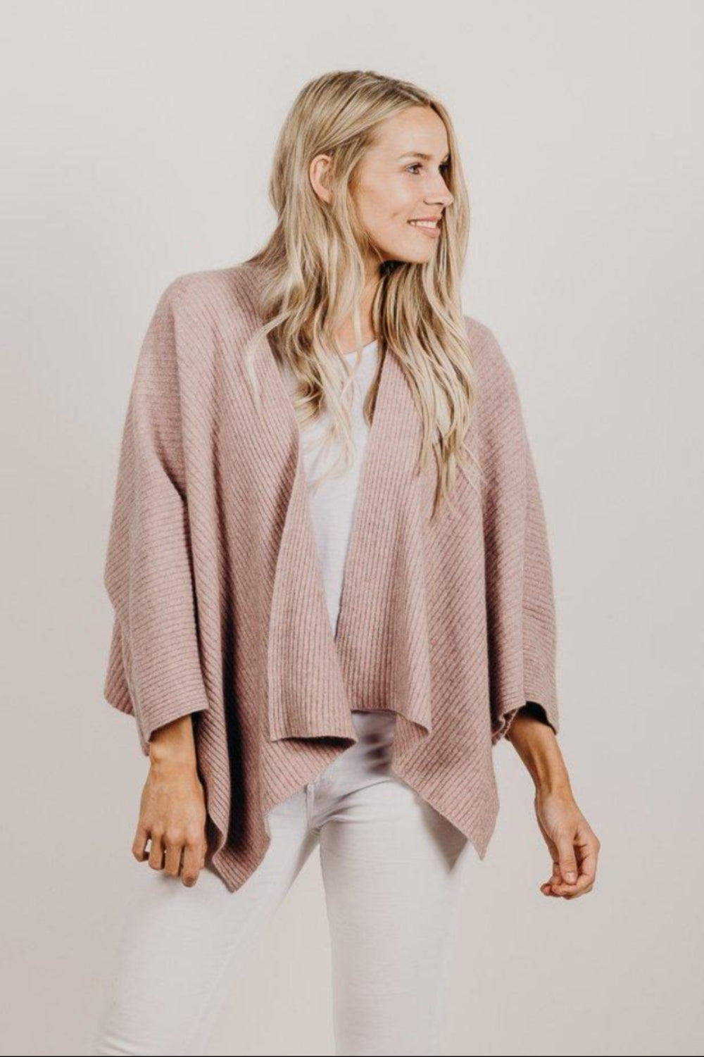 Woman wearing a pink knitted cardigan over a white top and pants on a plain background