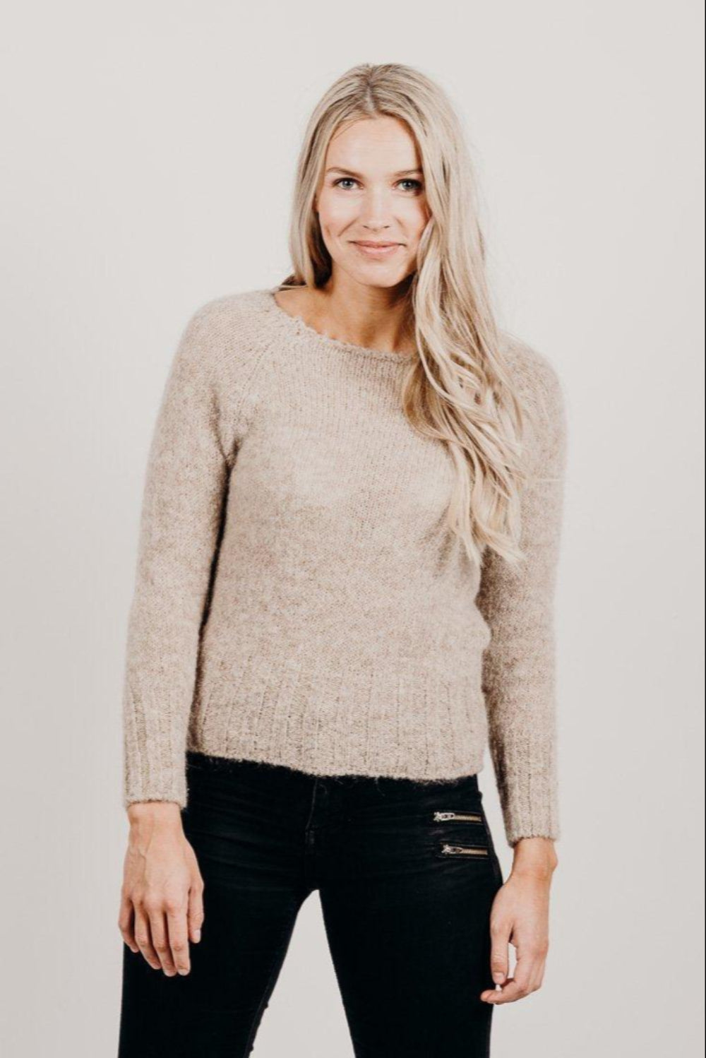 Woman wearing a beige sweater and black pants against a plain background