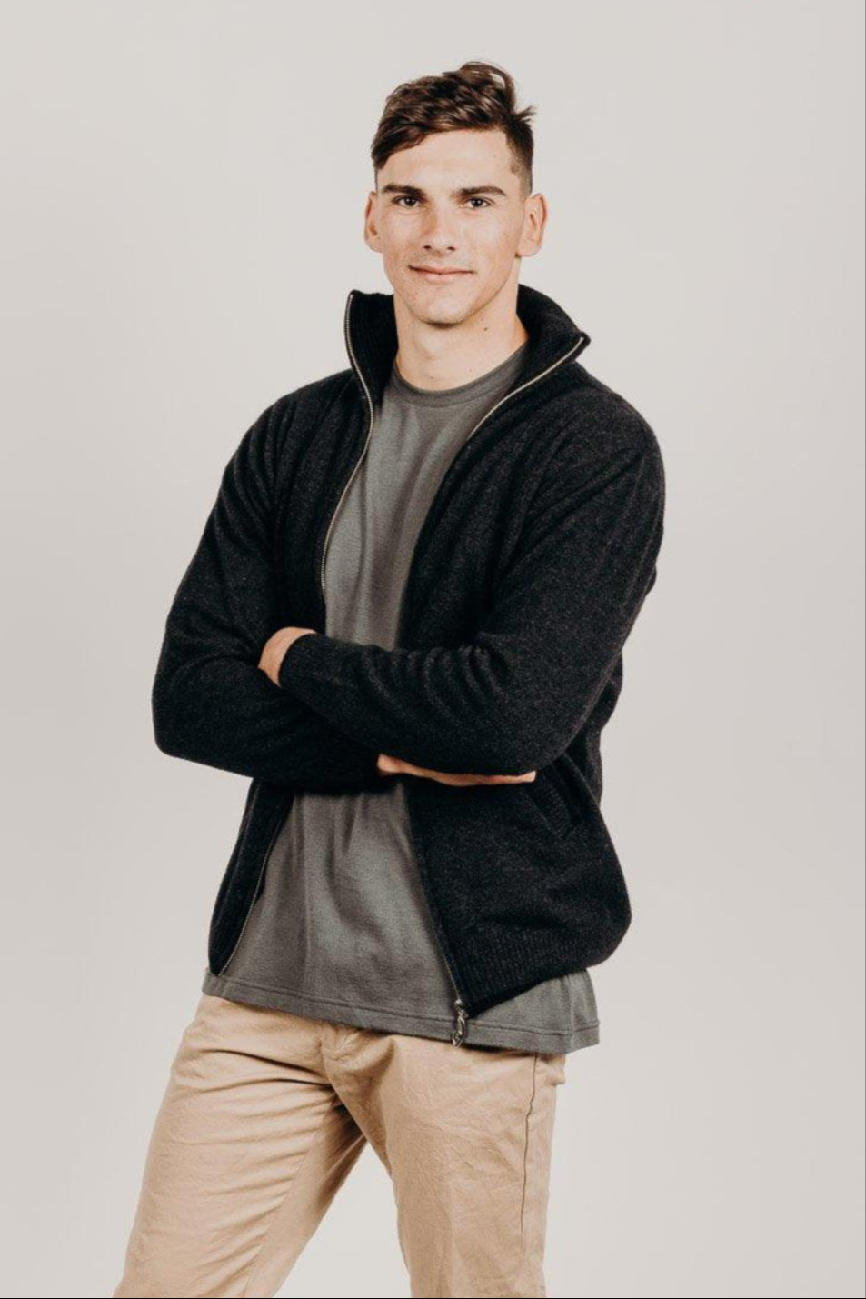 Man wearing a black  zip jacket over a gray shirt with beige pants on a plain background