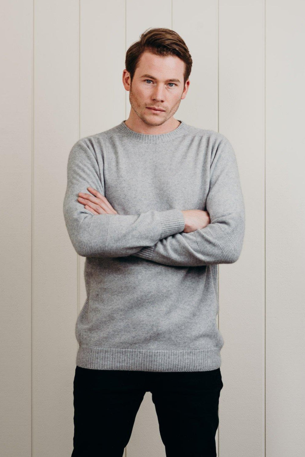 Man wearing a gray sweater with arms crossed against a light wooden panel background