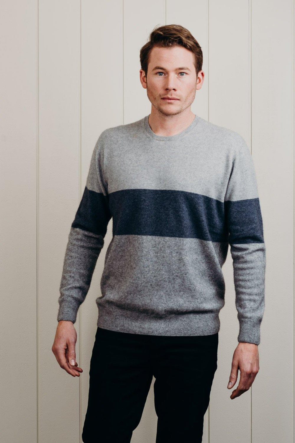 A man standing against a white background, wearing a gray and blue striped Merinosilk town sweater, paired with black pants.