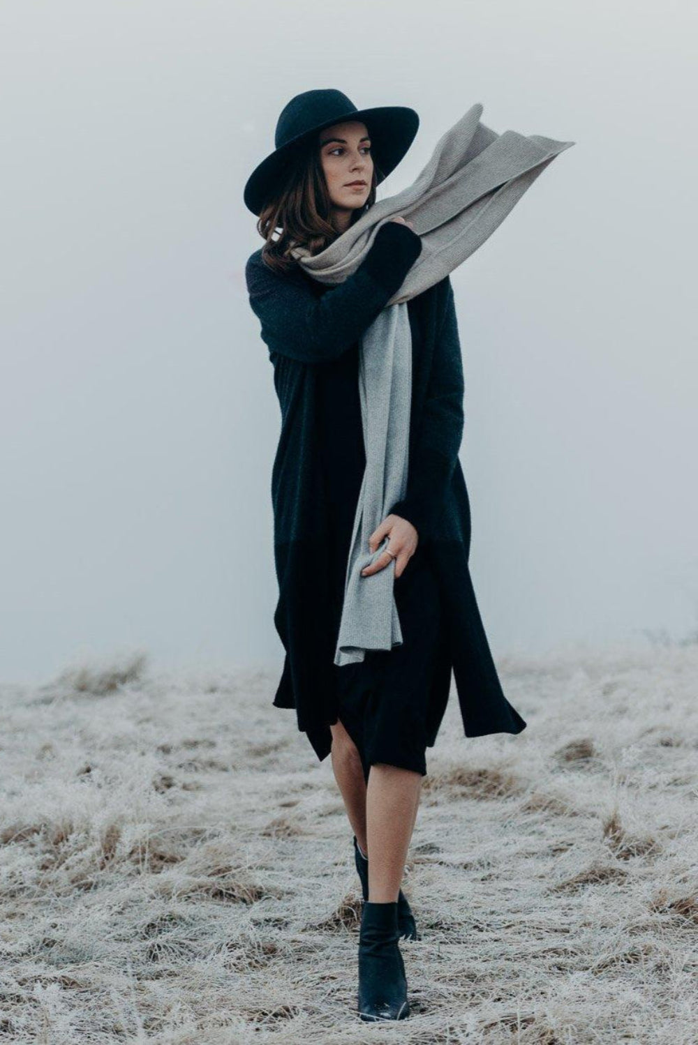 Person wearing a dark coat, hat, and scarf standing on a foggy field.