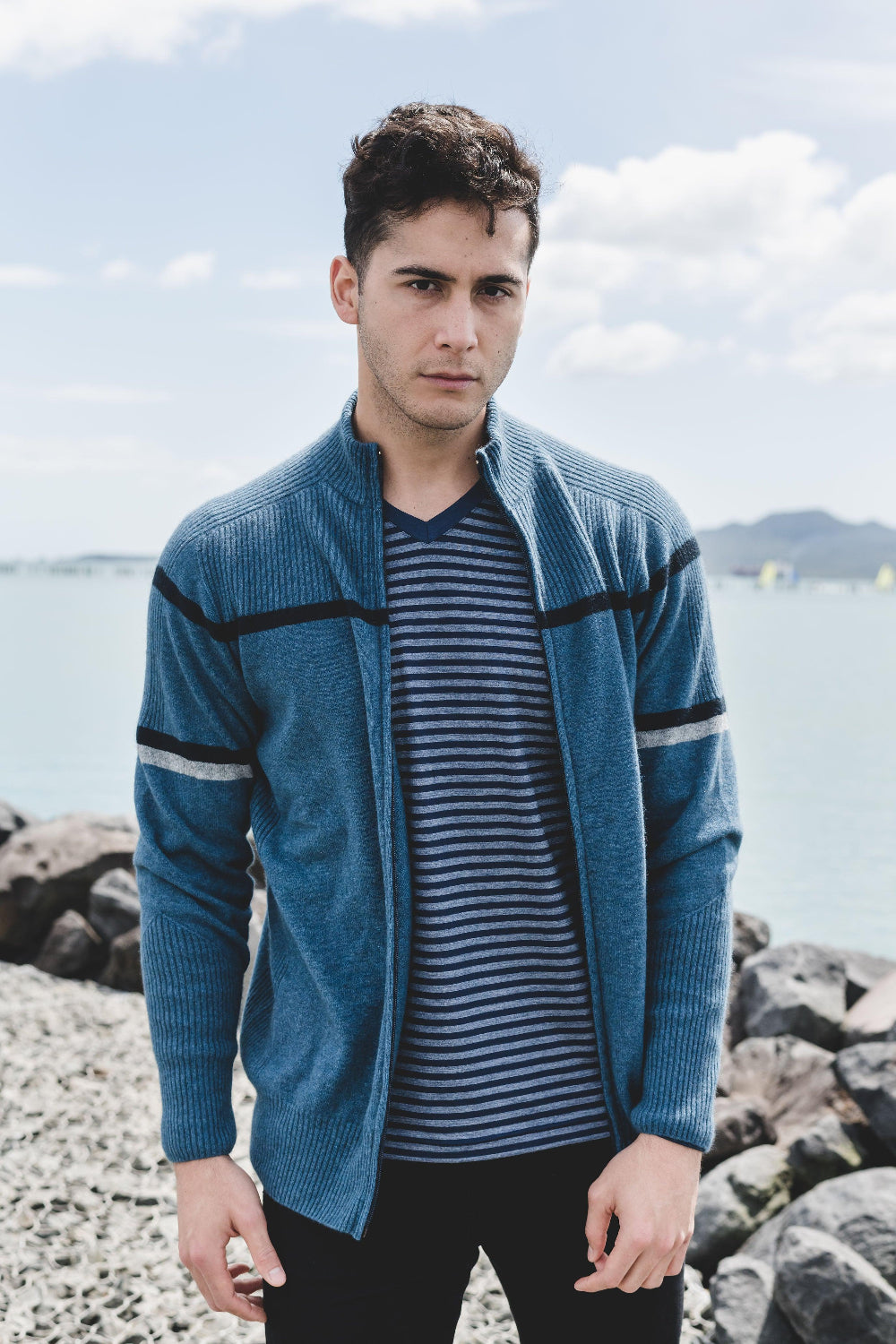 Man wearing a blue cardigan over a striped shirt by a body of water with rocks.