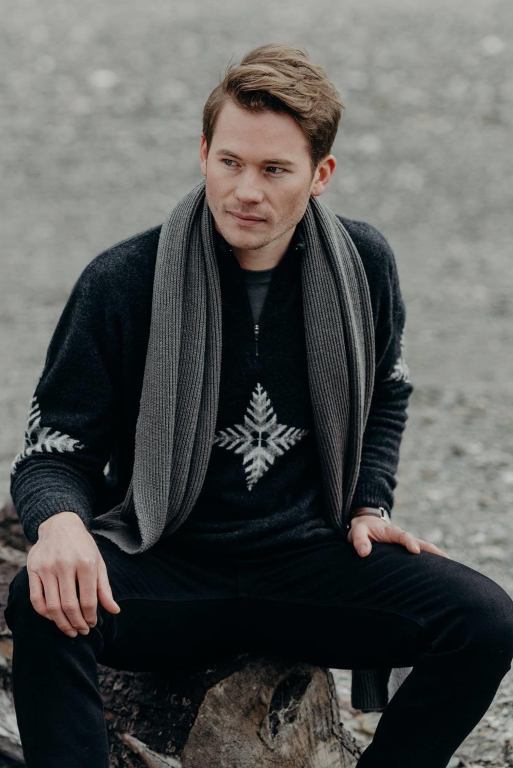 Man sitting on a log wearing a dark sweater with a patterned scarf and black pants.
