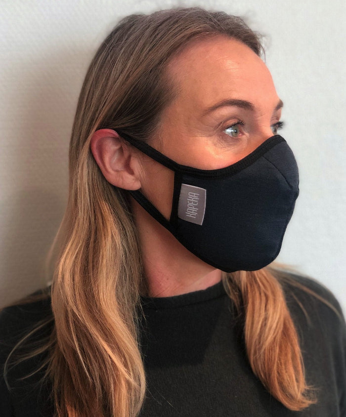Woman wearing a black travel mask with adjustable elastic straps, made from merino soft & comfort next to skin, with a label showing 'Made in New Zealand', and highlighting features such as 100% breathable, adjustable elastic straps, and a machine washable filter.