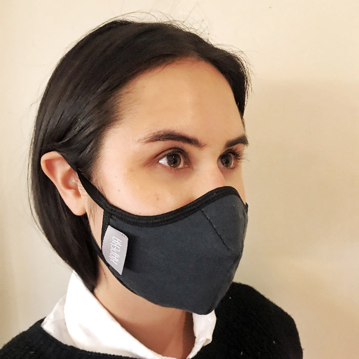 Woman wearing a black travel mask with adjustable elastic straps, made from merino soft &amp; comfort next to skin, with a label showing &#39;Made in New Zealand&#39;, and highlighting features such as 100% breathable, adjustable elastic straps, and a machine washable filter.