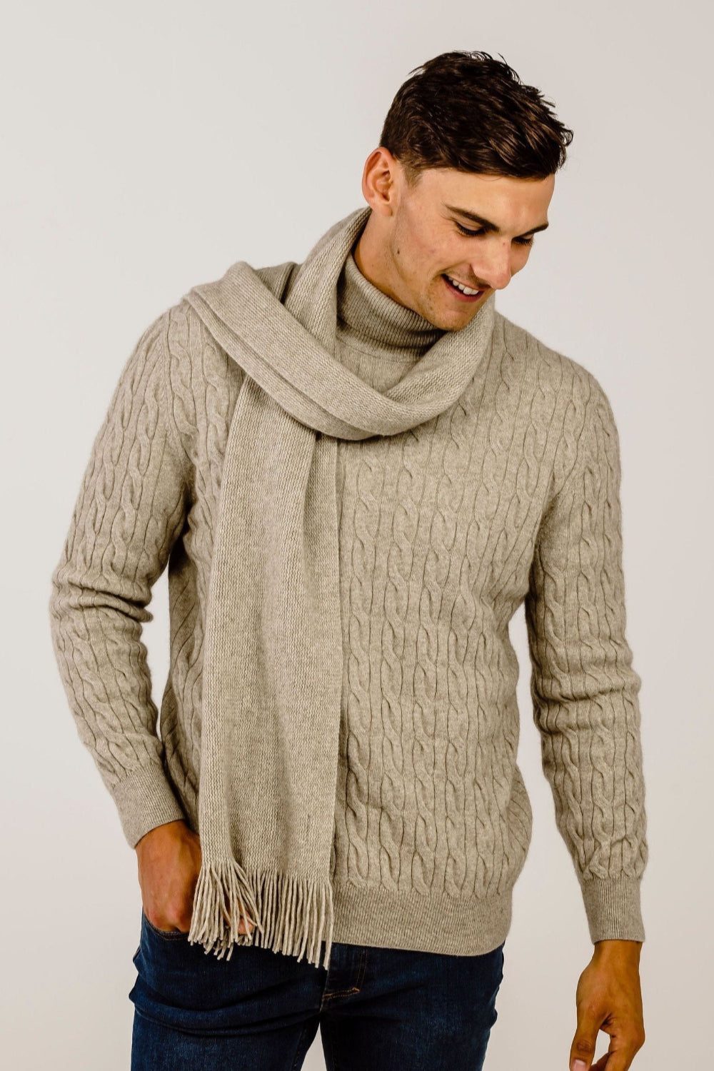 Man wearing a beige cable knit sweater and scarf against a plain background