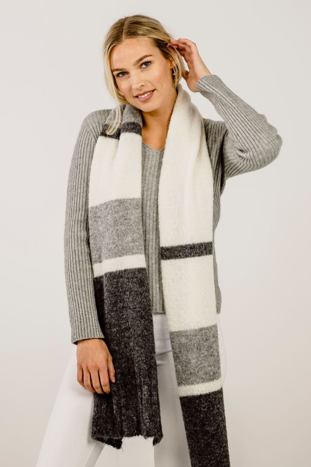 A woman wearing a gray and white striped alpaca scarf, paired with a gray sweater and white pants.