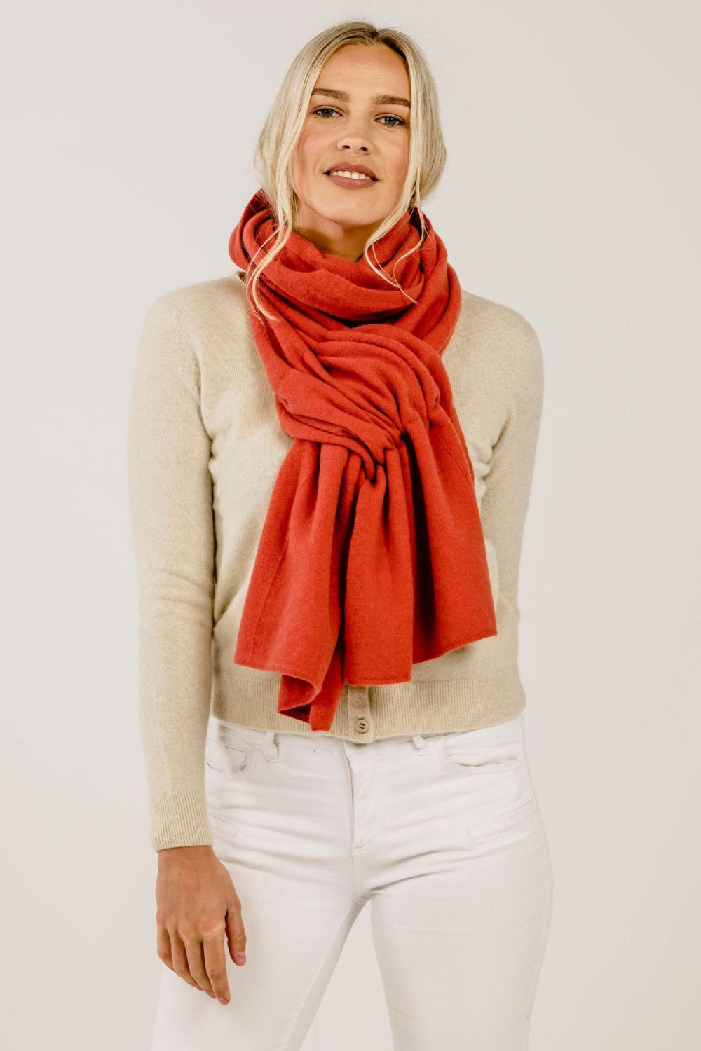 Woman wearing a red scarf, beige sweater, and white pants on a plain background