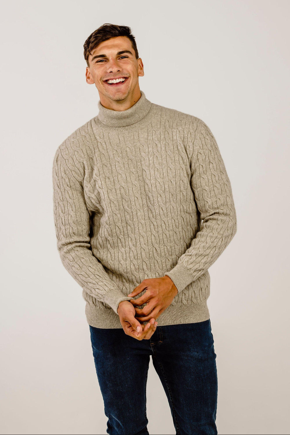 Man wearing a beige cable knit sweater and dark jeans against a plain background