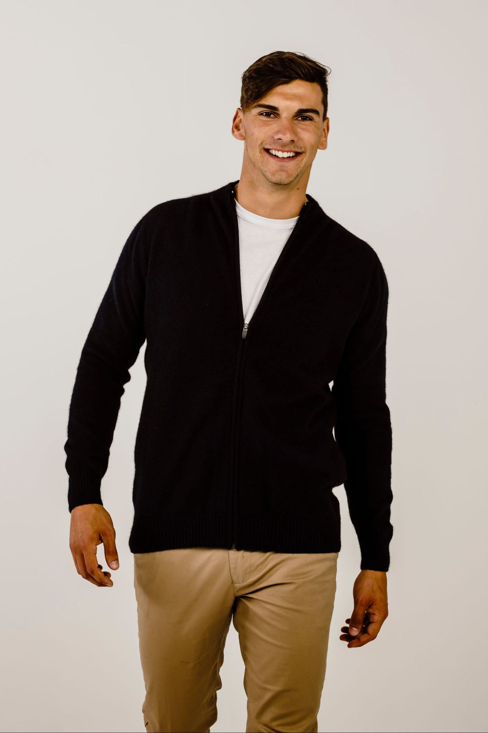 Man wearing a black cardigan over a white shirt and beige pants on a plain background