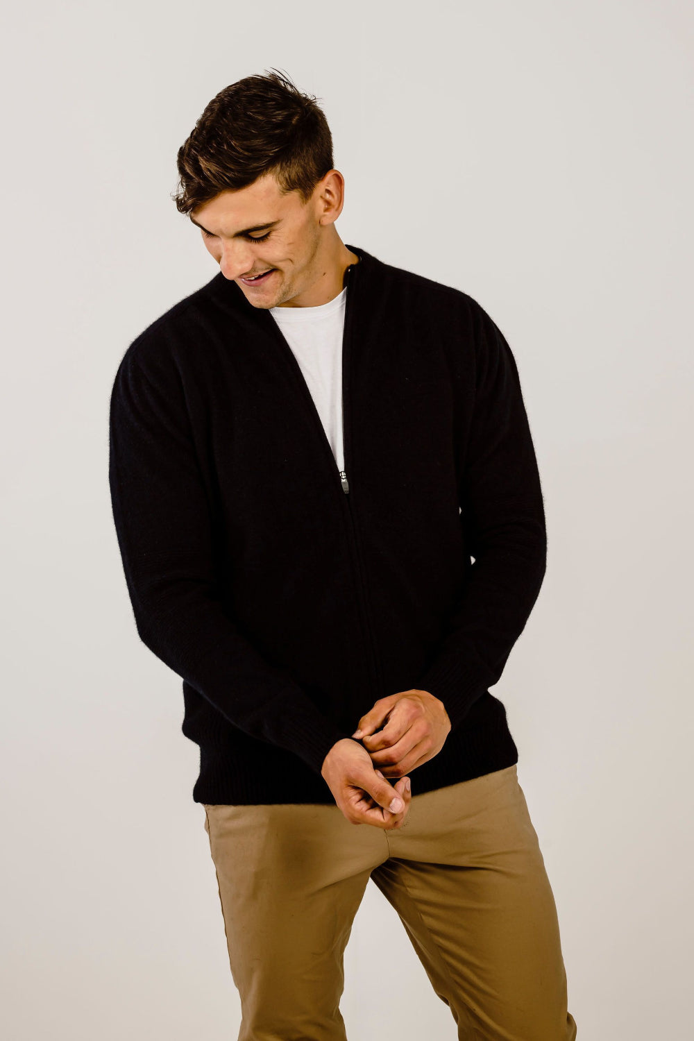 Man wearing a black sweater over a white shirt and beige pants on a light gray background
