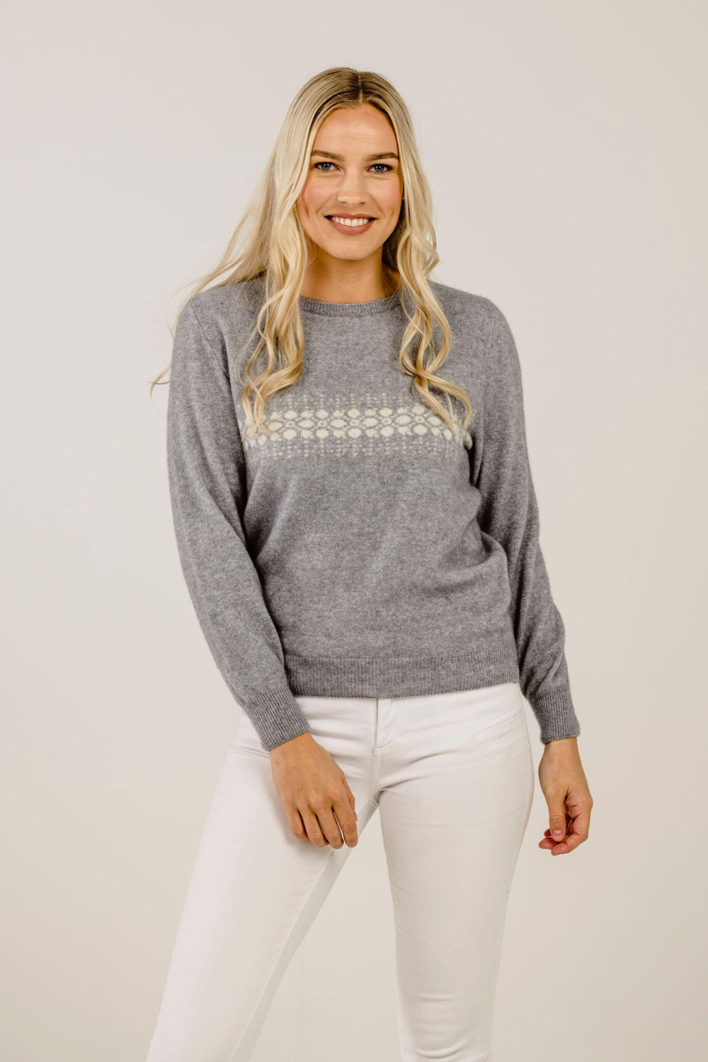 A woman standing, wearing a gray cashmere sweater with a crew neck and snow crystal design on the front, paired with white pants.