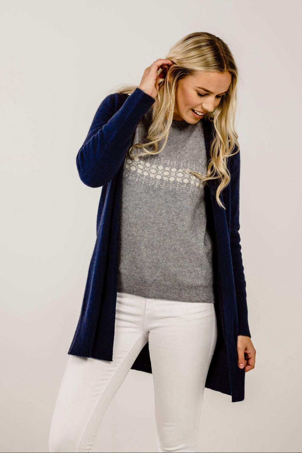 Woman wearing a navy cardigan, gray sweater, and white pants on a plain background