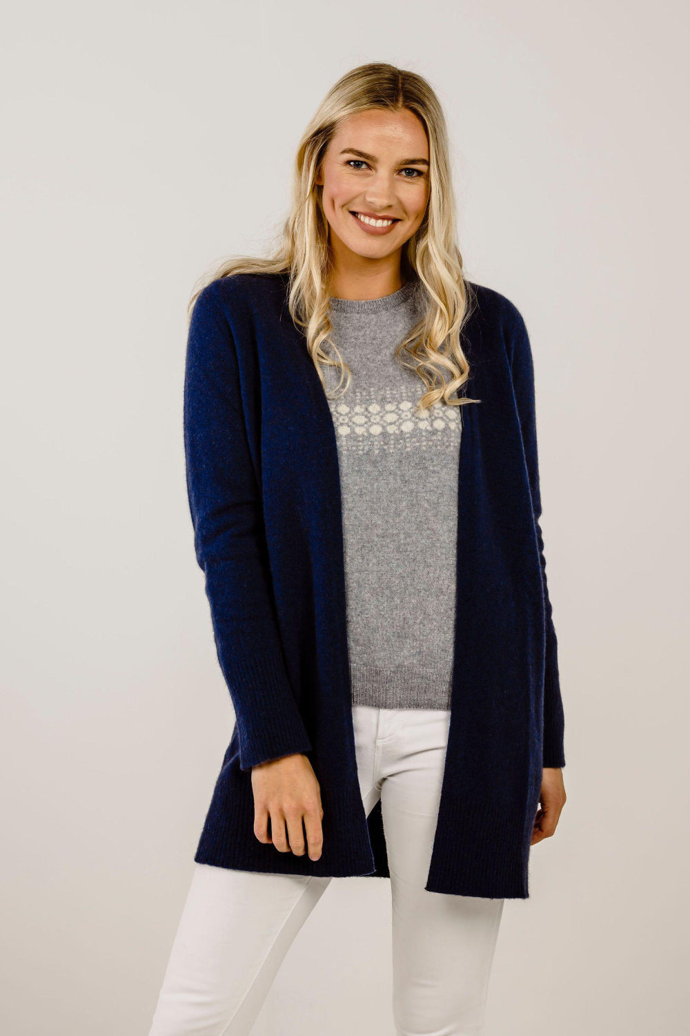 Woman wearing a navy cardigan over a gray sweater with white pants on a plain background