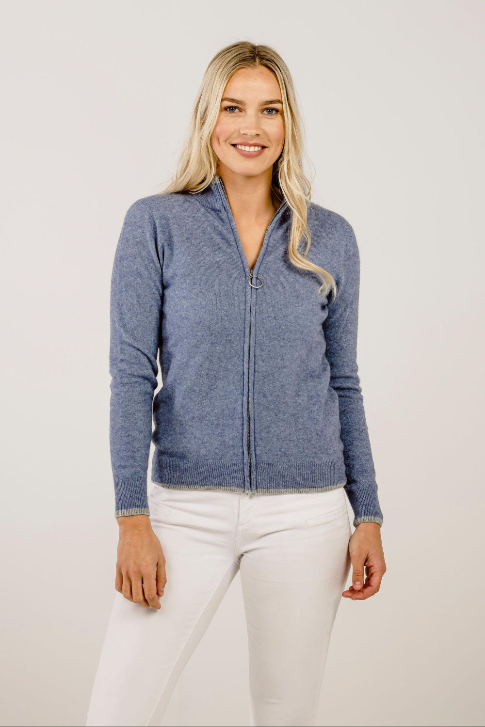 Woman wearing a blue zip-up sweater and white pants on a plain background
