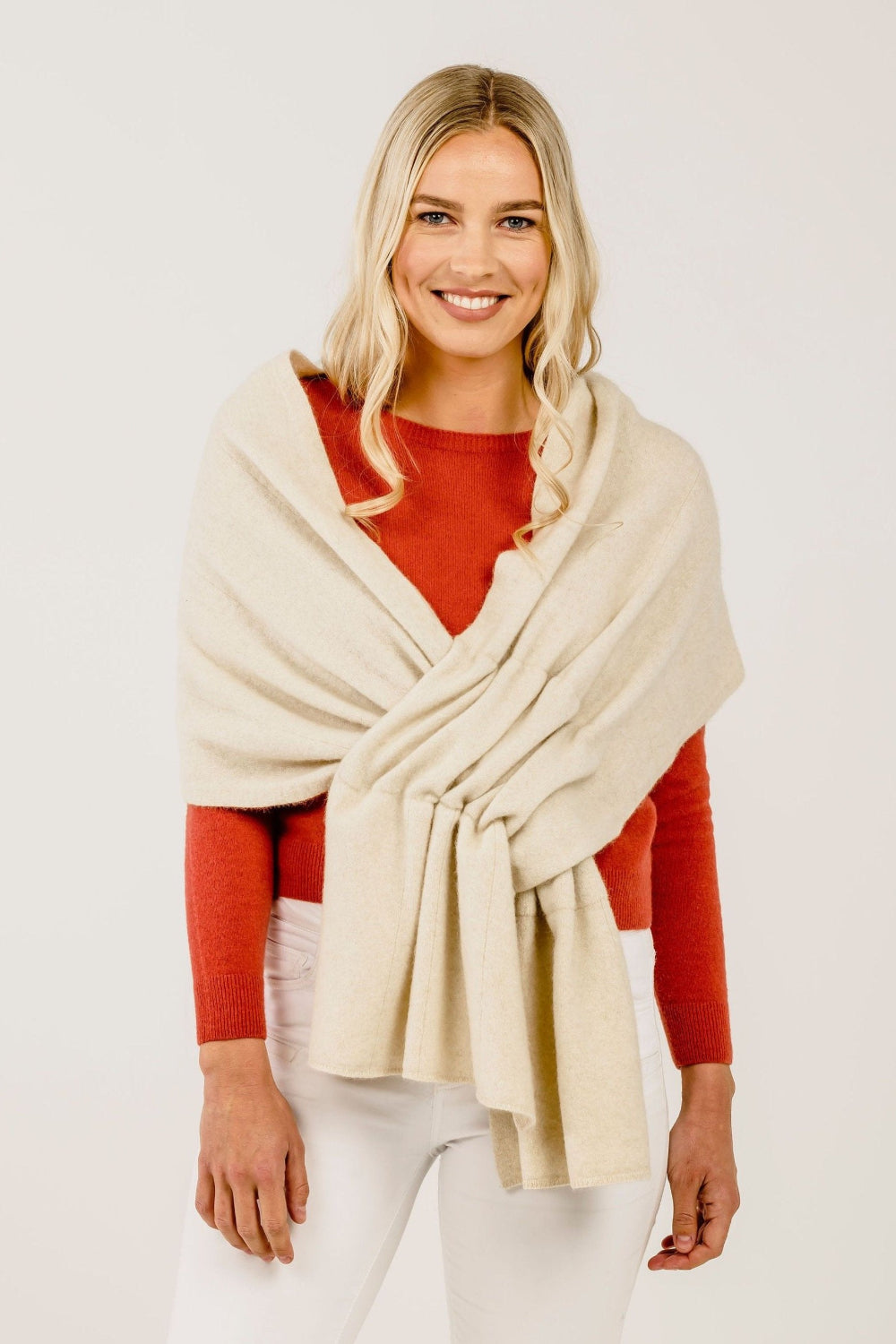 A woman wearing a cream-colored cashmere stole with a subtle texture, draped over her shoulders. She is also wearing a red top and white pants.