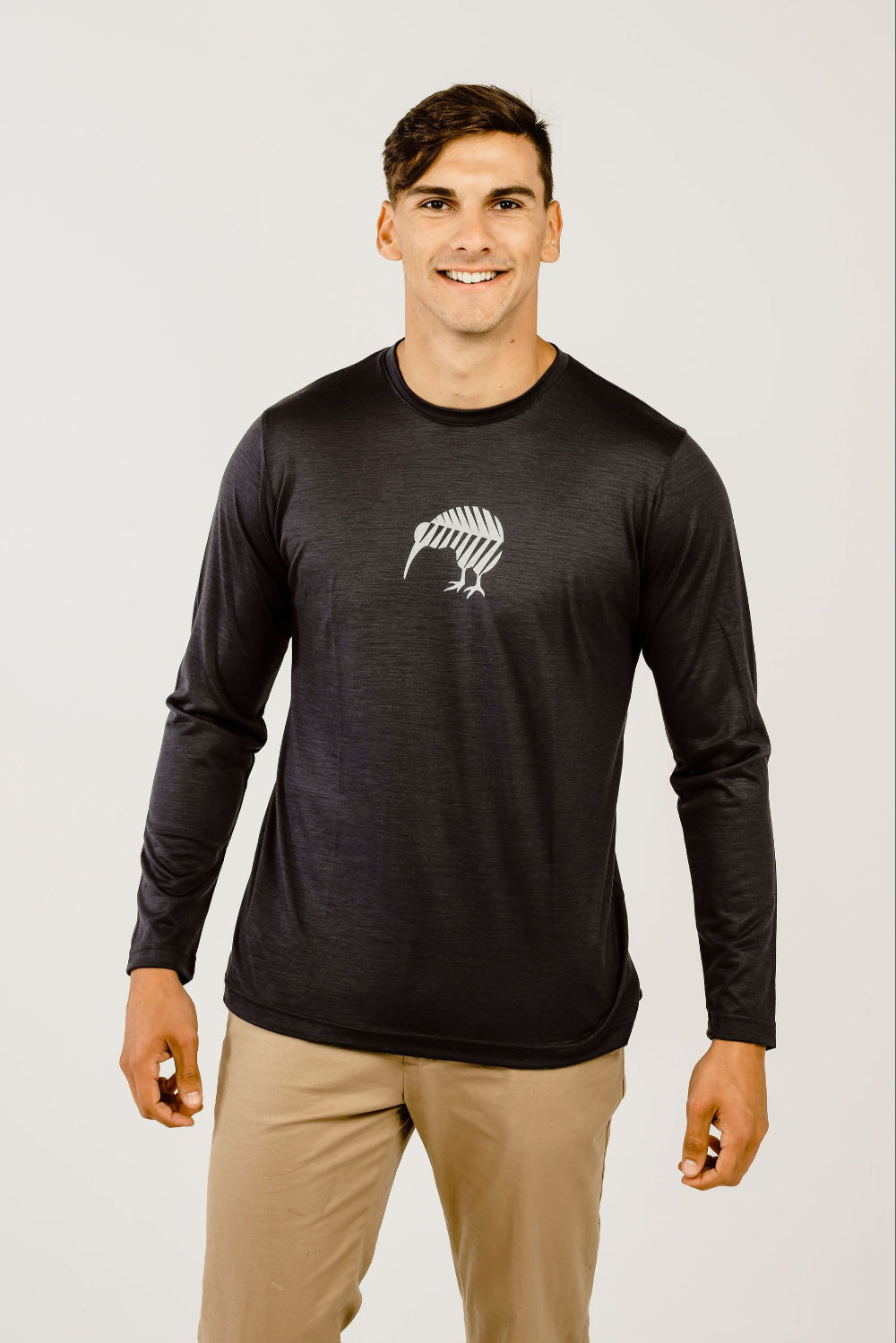 A male model wearing a long sleeve black Merino wool t-shirt with a white print of a Kiwi bird on the front and beige pants.