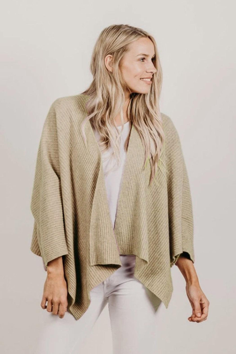 Woman wearing a beige knitted cardigan over a white top and pants on a plain background